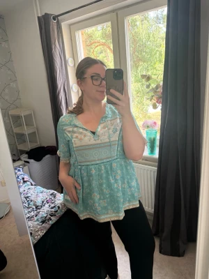 Turkos blommig boho-blus med knyt - Söt turkos blus med blommigt mönster och boho-vibbar. Blusen har v-ringning med knytband, lös passform och korta ärmar. Dekorativa spetsdetaljer och mönstrad kant nedtill. Perfekt för en avslappnad och trendig stil. Från märket Odd Molly i storlek 4 som enligt deras storlekguide motsvarar en storlek XL. Mycket fint skick, jag hittar inga synliga anmärkningar eller defekter. 