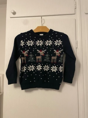 Mörkblå jultröja med renar Polarn O. Pyret - Mysig mörkblå stickad tröja från Polarn O. Pyret med vita snöflingor och renar med röda detaljer. Perfekt för vintern och julen. Rund halsringning och ribbade muddar. Passar barn i storlek 110 (4-5 år).