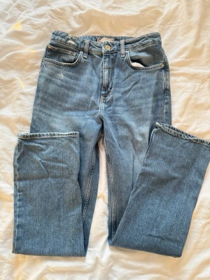 Blå raka jeans med hög midja - Säljer ett par klassiska blå jeans med rak passform och hög midja. Jeansen har fem fickor, bälteshällor och stängs med knapp och dragkedja. Materialet är kraftig denim i 100% bomull. Perfekta till en avslappnad och trendig look.