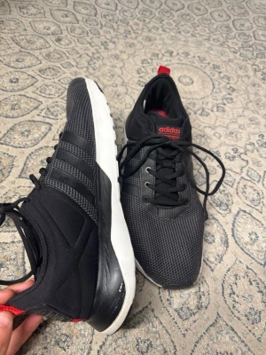 Svarta Adidas sneakers med röd detalj - Snygga svarta sneakers från Adidas med klassiska tre ränder på sidan, mesh-överdel och vita sulor. Skorna har röda detaljer på plösen och hälen samt svarta skosnören. Perfekta för en sportig och avslappnad stil. Ser ganska bra ut små tecken på att de är använda, Storlek 47 1/3, fraktar samma dag eller dagen efter :)