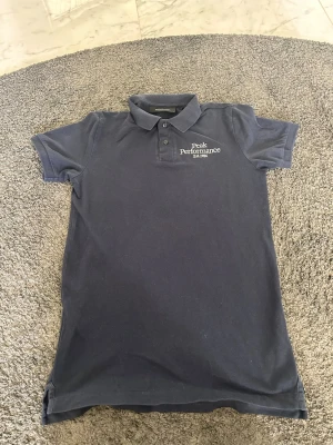 Marinblå pikétopp Peak Performance - Snygg marinblå pikétröja från Peak Performance med klassisk krage och knappar framtill. Diskret logga broderad på bröstet. Tillverkad i bomull för en skön känsla och stilren look. Perfekt till jeans eller shorts. Kontakta mig vid frågor!