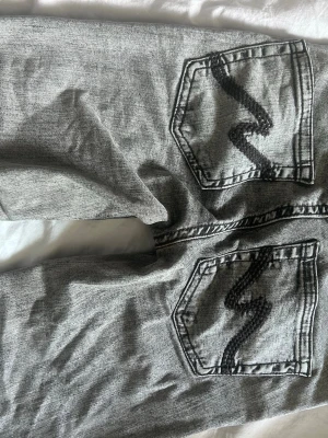 Grå jeans från Nelly - Säljer ett par gråa jeans från Nelly med raka ben och coola bakfickor med svart sicksack-detalj. Jeansen har klassisk femficksmodell och knappgylf. Perfekta för en avslappnad och trendig look.
