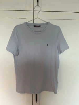 Morris T-Shirt - En stilren ljusblå t-shirt från Morris i bomull med rund hals och korta ärmar. På bröstet finns en liten broderad logga i svart som ger en clean look. Perfekt till jeans eller shorts för en enkel och snygg outfit.