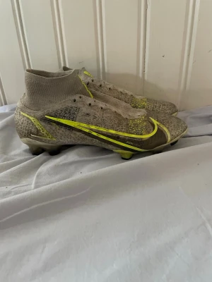 Nike Mercurial fotbollsskor beige/gul - Säljer ett par Nike Mercurial elite fotbollsskor i beige med gula detaljer och svart swoosh. Skorna har strumpliknande skaft för extra stöd och greppvänlig sula med dobbar, perfekta för spel på gräs. Tillverkade i syntetmaterial för lätt känsla och bra bollkontroll. (Köpte för 3400)