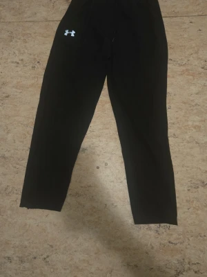 Svarta leggings från Under Armour - Säljer ett par svarta byxor från Under Armour med logga på vänster lår och reflekterande detaljer vid bensluten. De har hög midja och är tillverkade i ett stretchigt material som passar perfekt till träning eller chill.