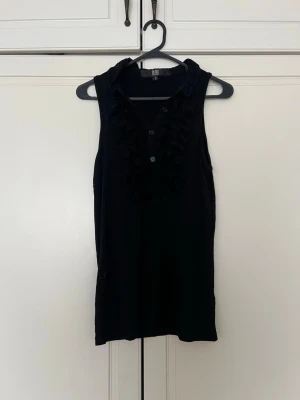 Svart ärmlös topp med volang från Bedo - Svart ärmlös topp från Bedo med volangdetaljer framtill och knappar vid halsen. Snygg och enkel modell som passar perfekt till jeans eller kjol. Gjord i mjukt material och har en klassisk, stilren look.