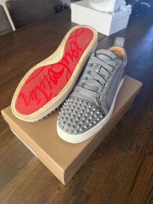 Louboutin skor - Säljer ett par gråa sneakers från Christian Louboutin i mocka med ikonisk röd sula. Skorna har coola nitar på tån och vita sulor. Perfekta för dig som vill sticka ut med en unik och lyxig stil. Klassisk låg modell med snörning. Storlek 40 men passar även 41 skulle jag säga. Allt på bilderna ingår hör av er vid funderingar😄✅