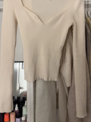 Vit ribbad långärmad topp - Säljer en vit ribbad topp med lång ärm och v-ringad hals. Toppen är croppad och har en mjuk, stretchig passform som sitter snyggt på kroppen. Perfekt att styla med jeans eller kjol för en clean look.