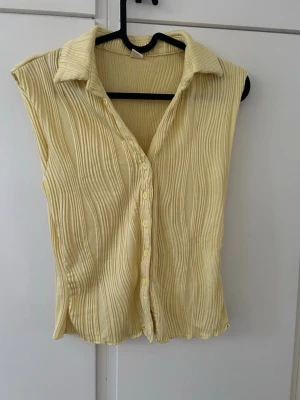 Gul ärmlös blus med krage - Säljer en ljusgul ärmlös blus med krage och knappar framtill. Blusen har ett plisserat tyg som ger en cool struktur och en avslappnad men ändå chic look. Perfekt att styla till jeans eller kjol för en trendig outfit.