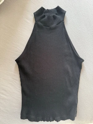 Svart ribbad halterneck topp NA-KD - Säljer en svart ribbad halterneck topp från NA-KD i storlek XS. Toppen har hög krage och är ärmlös med en croppad passform.  Aldrig använd