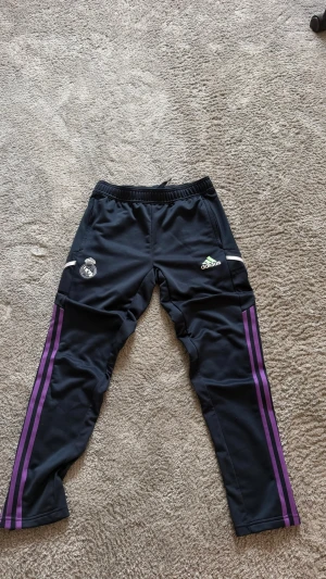 Svarta Adidas Real Madrid träningsbyxor - Säljer ett par svarta träningsbyxor från Adidas med Real Madrid-logga på vänster ben. Byxorna har lila ränder längs sidorna och vita detaljer. Tillverkade i ett sportigt, lätt material med dragkedjor vid fickorna. Perfekta för fotbollsträning eller gymmet.