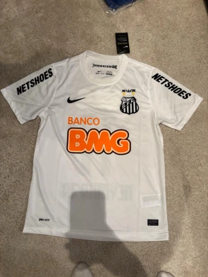 Santos retro tröja #11 Neymar jr - Säljer en vit Santos matchtröja från Nike med Neymar Jr och nummer 11 på ryggen. Tröjan har tryckta sponsorer som BMG och Netshoes, samt Santos klubbmärke på bröstet. Tillverkad i lätt och ventilerande Dri-FIT-material, perfekt för fotboll eller träning.