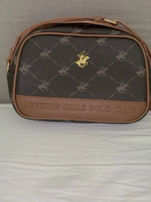 Brun axelremsväska Beverly Hills Polo Club - Snygg brun axelremsväska från Beverly Hills Polo Club med guldfärgad logga framtill. Väskan har ett mönster med klubbens ikoniska polospelare och justerbar axelrem i brunt tyg. Materialet är konstläder och väskan har en klassisk, rundad form.
