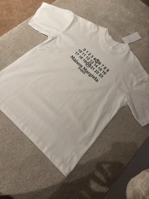 Vit Maison Margiela t-shirt - Vit t-shirt från Maison Margiela med det ikoniska numrerade trycket på bröstet och texten 'Maison Margiela Paris' i svart. Klassisk rund hals och rak passform. Perfekt för dig som gillar minimalistisk och stilren design.