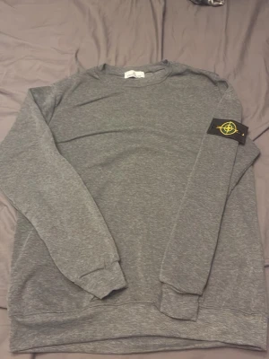 Grå sweatshirt från Stone Island - Säljer en stilren grå sweatshirt från Stone Island med klassisk rund hals och den ikoniska loggan på vänster ärm. Tröjan har långa ärmar och ribbade muddar vid ärmslut och nederkant. Perfekt för en avslappnad och trendig look.