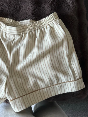 Randiga beige shorts från NA-KD - Säljer ett par snygga beige shorts från NA-KD med tunna bruna ränder. De har elastisk midja och är gjorda i ett mjukt bomullsmaterial. Perfekta för en avslappnad och trendig look under varma dagar. Aldrig använt 