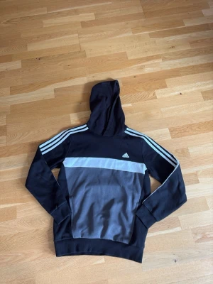 Svart och grå hoodie från Adidas - Snygg hoodie från Adidas i svart och grått med klassiska vita ränder på ärmarna och Adidas-logga på bröstet. Tröjan har huva och muddar vid ärmslut och nederkant. Perfekt för en sportig och avslappnad stil.