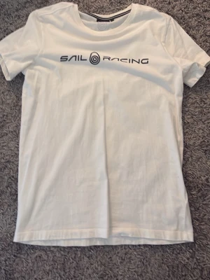 Vit t-shirt från Sail Racing - Vit t-shirt från Sail Racing med tryckt logga över bröstet. Klassisk rund halsringning och korta ärmar. Tillverkad i mjuk bomull för en skön känsla. Perfekt till jeans eller shorts för en clean look.