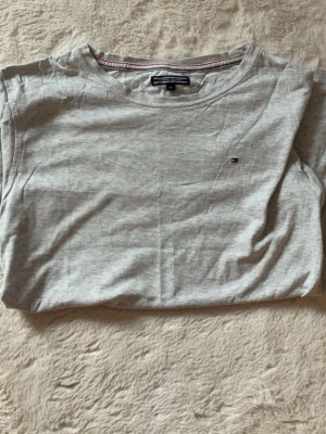 Ljusgrå t-shirt från Tommy Hilfiger - Snygg ljusgrå t-shirt från Tommy Hilfiger med rund halsringning och diskret logga broderad på bröstet. Tillverkad i mjukt bomullsmaterial som känns skönt mot huden. Perfekt basplagg till garderoben.