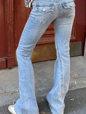 Ljusblåa lågmidjade true religion jeans - Säljer ett par ljusblå bootcut jeans med snygga fickdetaljer bak och klassisk femficksmodell. Jeansen har utsvängda ben och cool tvättad look. Perfekta för dig som gillar retrovibbar och vill ha ett par jeans som sticker ut lite extra.