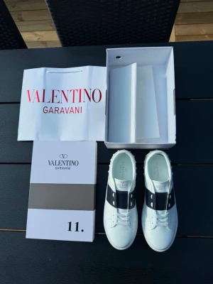 Valentino Garavani vita sneakers - Stilrena vita sneakers från Valentino Garavani med svart band och nitar på sidan. Skorna har en klassisk låg profil, rund tå och är tillverkade i slätt läder. Sulan har ett rutmönstrat grepp och logga på hälen. Perfekta för dig som gillar exklusiv och modern design.