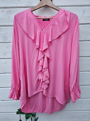 Rosa blus med volangdetaljer L - Snygg rosa blus med långa ärmar och volangdetaljer längs V-ringningen och framtill. Blusen har en lös passform och bred volangkrage som ger en romantisk vibe. Perfekt att styla med jeans eller kjol för en trendig look.