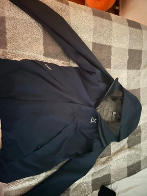 Mörkblå regnjacka från Haglöfs - Snygg mörkblå regnjacka från Haglöfs med huva och dragkedja framtill. Jackan är tillverkad i ett tunt, vattentätt material och har meshfoder på insidan för extra komfort. Perfekt för regniga dagar och outdoor-äventyr.