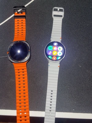 Säljer båda  - Två Samsung smartklockor med rund urtavla och silikonband. Den ena har ett orange band och svart boett, den andra har ett vitt band och silverfärgad boett. Båda har touchskärm och moderna funktioner för appar och notiser.