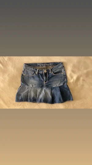 Blå volang jeanskjol - Säljer en blå jeanskjol med volangkant och dekorativa sömmar. Kjolen har klassiska fickor fram och bak samt dragkedja och knapp framtill. Perfekt för dig som gillar en avslappnad men trendig stil.