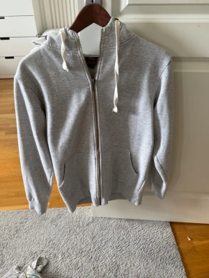 Ljusgrå zip hoodie med luva - Säljer en ljusgrå hoodie med dragkedja och luva. Tröjan har två fickor framtill och vita dragsnören i huvan. Tillverkad i mjukt sweatshirtmaterial, perfekt för chill dagar eller när du vill ha något bekvämt.