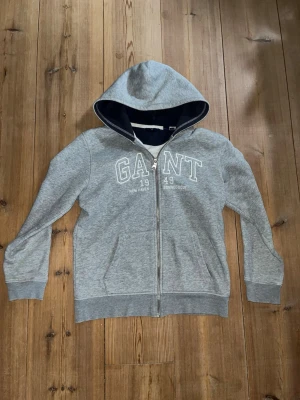 Grå zip-hoodie från GANT - Snygg grå hoodie från GANT med dragkedja och vit broderad logga framtill. Mörkblått innerfoder i huvan och mjukt material på insidan. Klassisk passform med ribbade muddar och fickor på sidorna. Perfekt för en avslappnad och chill stil!Helt nyskick och aldrig använd!      Nypris:1449kr.    Mitt pris:699