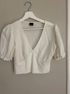 Vit croppad omlottopp - Säljer en vit croppad topp med omlottdesign och puffiga korta ärmar. Toppen har v-ringning och resår vid ärmsluten för en söt look. Perfekt att styla med höga jeans eller kjol.