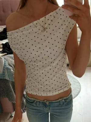 Vit offshoulder topp med prickar - Säljer en vit offshoulder topp med svarta prickar. Toppen har en snygg draperad axel och sitter tight mot kroppen. Perfekt för dig som gillar trendiga detaljer och vill sticka ut lite extra.