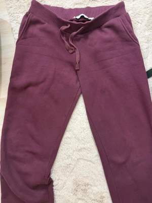 Vinröda mjukisbyxor med dragsko - Säljer ett par vinröda mjukisbyxor med resår i midjan och dragsko. Byxorna har två sidofickor och är gjorda i mjukt, bekvämt material som passar perfekt för chill dagar. Klassisk rak passform och ribbade muddar vid midjan. Några fläckar på ena benet