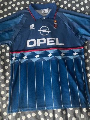 Blå AC Milan retro fotbollströja - Säljer en blå retro AC Milan fotbollströja från Lotto med Opel-logga på bröstet. Tröjan har klassiskt krage, röda och vita detaljer samt klubbmärke och stjärna på bröstet. Snyggt mönster och tunnt, sportigt material. Perfekt för samlare eller fans!