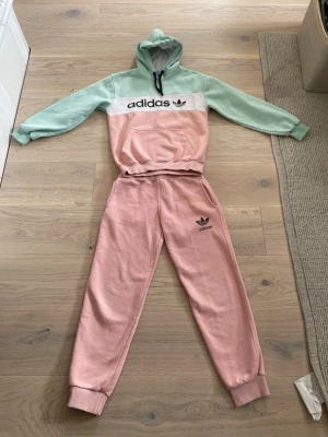 Adidas pastellfärgat loungewear-set - Man Kan förhandlas. kommer från ett djur frit hemMysigt loungewear-set från Adidas med hoodie och byxor. Tröjan har mintgröna ärmar, rosa kropp och vit panel med svart Adidas-logga. Byxorna är ljusrosa med Adidas-tryck på benet. Setet är gjort i mjuk bomullsmix och har avslappnad passform med muddar.