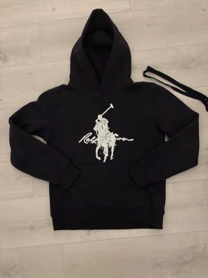 Ralph lauren hoodie  - Säljer en mörkblå hoodie från Ralph Lauren med stor vit Polo-logga och ryttare på bröstet. Hoodien har huva och en klassisk känguruficka framtill. Snöret till huvan medföljer löst. Perfekt för en avslappnad och stilren look. Snöret åker med på sidan då jag tagit bort de innan för att jag tyckt det varit för långt och bara varit i vägen. Tröjan sitter som S/M och även fast tröjan är i storlek M. Vid fundringar och frågor så är det bara att skriva ✍️!