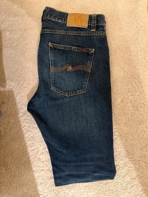 Nudie Jeans - Säljer ett par klassiska Lean Dean Deep Ocean  från Nudie Jeans (W31L30) med snygga kontrastsömmar och vågig detalj på bakfickan. Jeansen har slim passform och är tillverkade i slitstark denim. Perfekta för en avslappnad och stilren look.