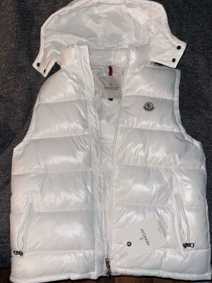Vit Moncler väst - Snygg vit dunväst från Moncler med avtagbar huva och två dragkedjefickor framtill. Västen har Moncler-logga på bröstet och quiltad design. Tillverkad i glansigt material med innerfoder och klassisk Moncler-serietikett på insidan.