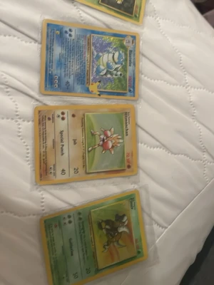 Pokémonkort Hitmonchan, Pinsir, Blastoise - Tre Pokémonkort: Hitmonchan med beige bakgrund, Pinsir med grön bakgrund och Blastoise med blå bakgrund. Alla korten är i plastfickor och har klassisk gul ram. Perfekt för samlare eller nostalgitripp!