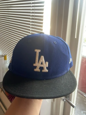 Los Angeles Dodgers Cap - This is a blue and black fitted cap with the iconic 'LA' logo on the front and 'Los Dodgers' embroidered on the side. The cap is likely an official New Era product, commonly associated with the Los Angeles Dodgers baseball team.