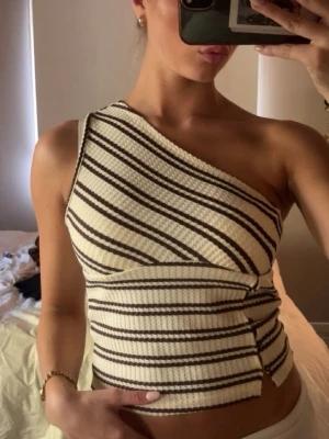 Randig oneshoulder topp i ribbad bomull - Snygg oneshoulder topp med diagonala bruna och vita ränder. Toppen är ribbad och har en tight passform som framhäver figuren. Perfekt för dig som vill sticka ut med en unik och trendig look.
