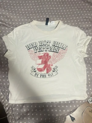 Vit Red Hot Chili Peppers t-shirt - Säljer en vit t-shirt från H&M Divided med Red Hot Chili Peppers-tryck och vingar i rött och svart. T-shirten har korta ärmar och rund hals. Perfekt för dig som gillar bandmerch och vill ha en avslappnad stil.