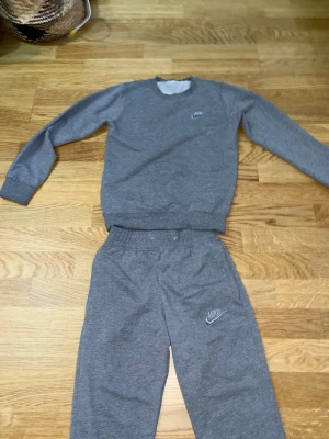 Grått set från Nike med sweatshirt - Säljer ett grått set från Nike med sweatshirt och matchande byxor. Båda delarna har Nike-logga tryckt på bröstet och benet. Setet är gjort i mjukt och bekvämt material, perfekt för chill dagar eller träning. Klassisk rund halsringning och resår i midjan på byxorna.