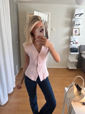Ljusrosa figursydd blus med knappar - Supersnygg ljusrosa blus med v-ringning och knappar framtill. Figursydd modell som framhäver midjan och har breda axelband. Perfekt till jeans eller kjol för en trendig look. Materialet känns mjukt och lätt, troligen bomull eller polyester.aldrig använd, lapp kvar 
