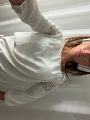 Vit blus med spetsdetaljer - Säljer en söt vit blus med fina spetsdetaljer på axlar och ärmar. Blusen har 3/4-ärmar och ett lätt, luftigt tyg – perfekt till både vardag och fest! Använd men i fint skick. Passar storlek S
