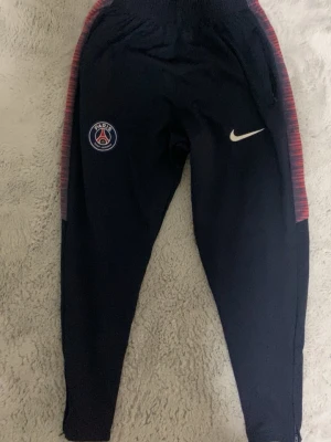 PSG träningsbyxor från Nike - Mörkblå träningsbyxor från Nike med PSG-logga på ena benet och vit Nike Swoosh på andra. Sidorna har röda och blå mönstrade ränder. Byxorna har smal passform och resår i midjan, perfekta för fotbollsträning eller chill. (Väldigt stretchiga) 