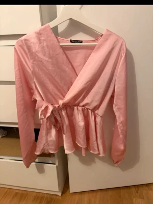 Rosa omlottblus från Missäy - Söt rosa omlottblus från Missäy med volang nedtill och knytband i midjan. Blusen har långa ärmar och en lätt glansig finish som ger en fräsch look. Perfekt att styla med jeans eller kjol för en trendig outfit.