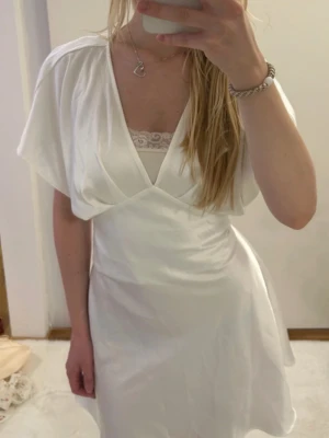 Vit omlottklänning med v-ringning - Säljer en vit omlottklänning med v-ringning och korta ärmar. Klänningen har en mjuk, svepande form och är tillverkad i ett glansigt material som ger en elegant look. Perfekt för sommarens alla tillfällen!