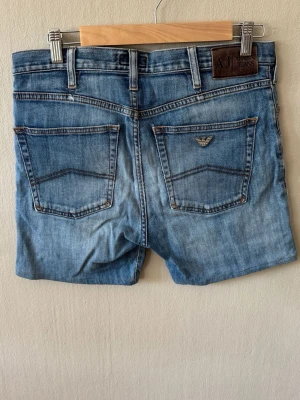 Blå jeansshorts från Armani Jeans - Säljer ett par blå jeansshorts från Armani Jeans med klassisk femficksdesign och ljus tvätt. Shortsen har bakfickor, varav en med metallogo, och en patch med logga i midjan. Perfekta för en avslappnad och trendig stil.0110